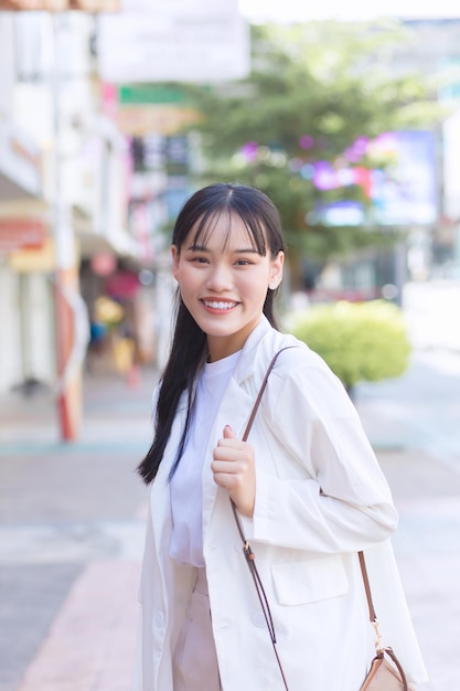 Confident young Asian female who wears white shirt, shoulder bag smiles happily while she is walking