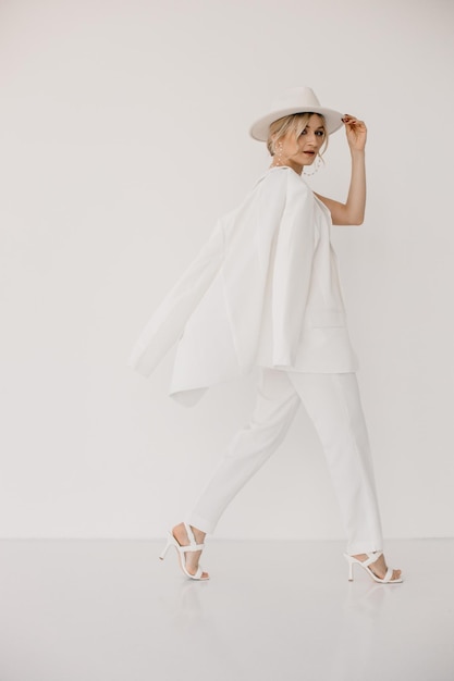 Confident young blond woman smiling looking at camera isolated on white background Studio portrait of successful friendly female in white suit and hat posing over white wall