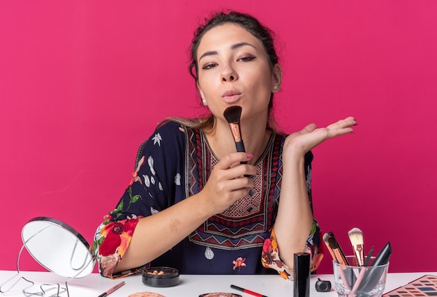 Confident young brunette girl sitting at table with makeup tools holding makeup brush isolated on pink wall with copy space