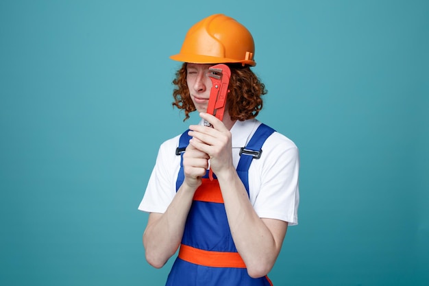 Confident young builder man in uniform holding gas wrench isolated on blue background