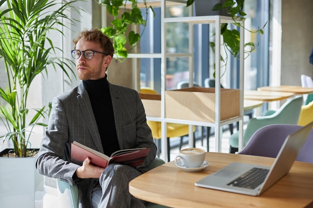 Confident young business coach holds a book and looks away sitting at a table with laptop in office