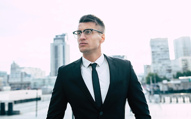 Confident young business man in formal wear and glasses standing outdoors with cityscape in the background