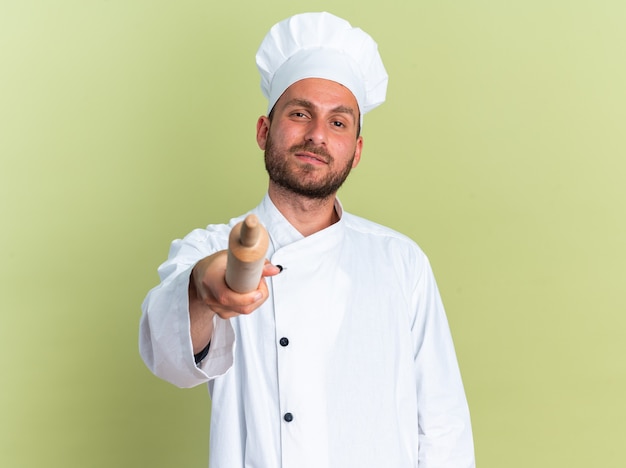 Confident young caucasian male cook in chef uniform and cap looking at camera pointing at camera with rolling pin isolated on olive green wall