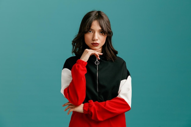 Confident young caucasian woman keeping hand under elbow and another hand under chin looking at camera isolated on blue background