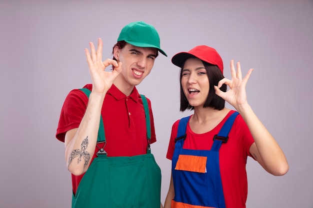 Confident young couple in construction worker uniform and cap looking at camera doing ok sign girl winking isolated on white wall