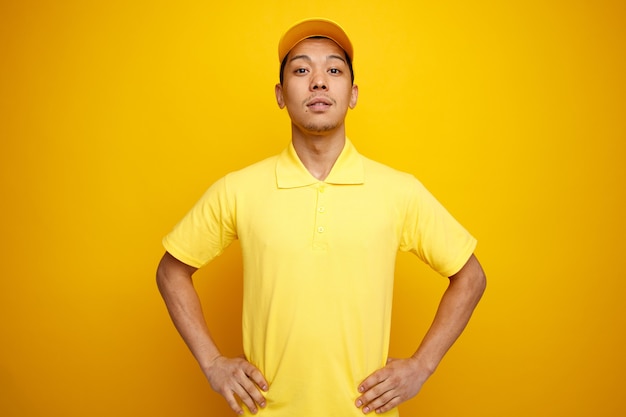 Confident young delivery man wearing cap and uniform keeping hands on waist 