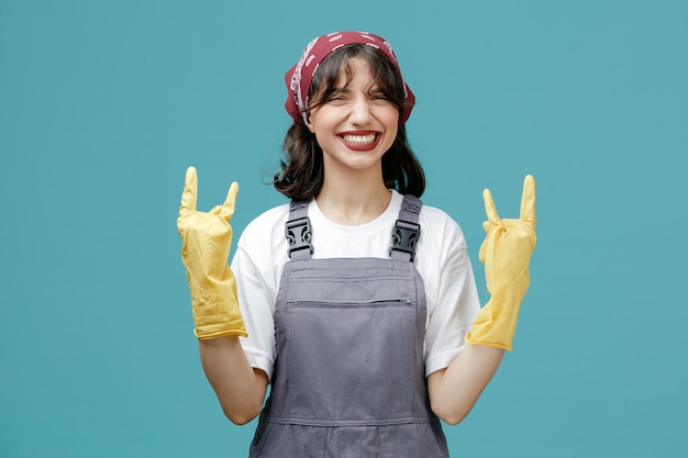 Confident young female cleaner wearing uniform bandana and rubber gloves looking at camera showing rock sign with both hands isolated on blue background