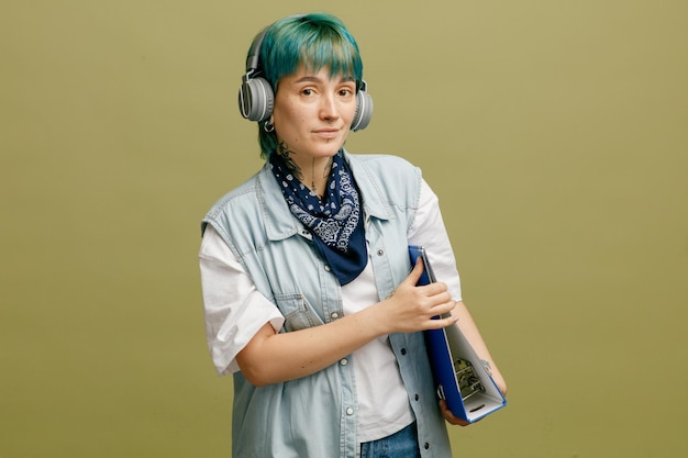 Confident young female student wearing headphones and bandana on neck holding folder with both hands looking at camera isolated on olive green background