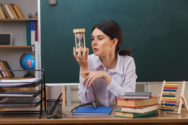 confident young female teacher holding and looking at sand watch sitting at desk with school tools in classroom