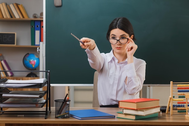 confident young female teacher wearing glasses points at side with pointer sitting at desk with school tools in classroom