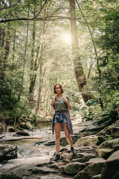 Confident young female traveler with backpack walking by the stones along the mountain river