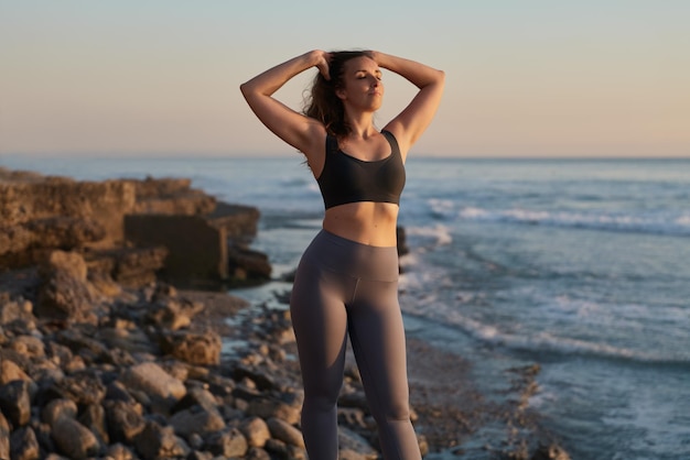 Confident young fit female touching hair on rocky seashore