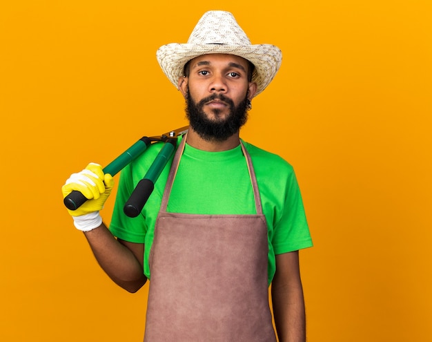 Confident young gardener afro-american guy wearing gardening hat and gloves putting clippers on shoulder 