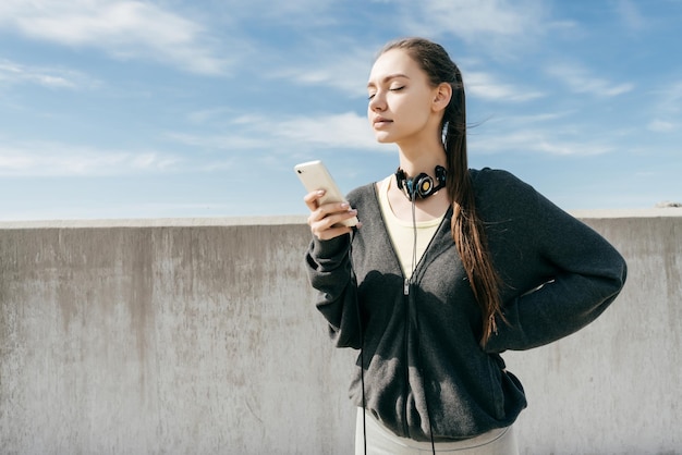 Confident young girl in black sports clothes went to a workout in the open air looks into her smartphone