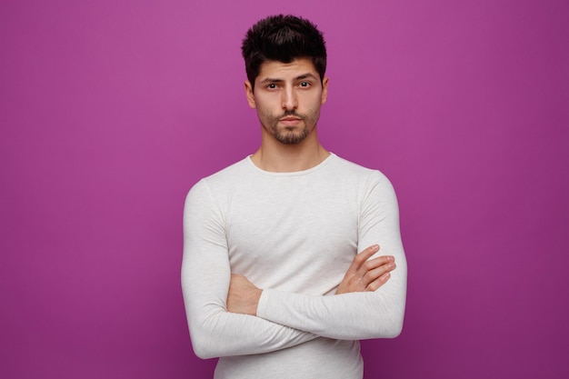 Confident young handsome man looking at camera with crossed arms on purple background