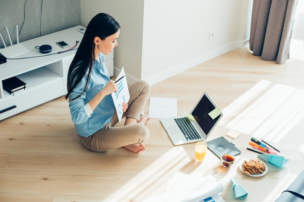 Confident young lady sitting on the floor in front of a laptop and showing graphics to the web camera