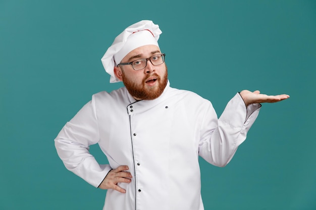 Confident young male chef wearing glasses uniform and cap looking at camera showing empty hand keeping another hand on waist isolated on blue background