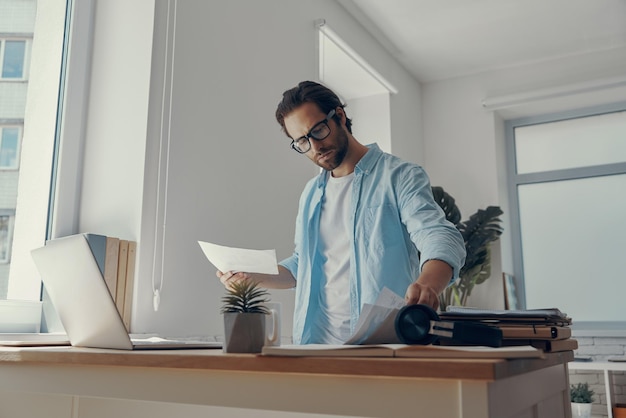 Confident young man doing some paperwork while standing near his working place in office