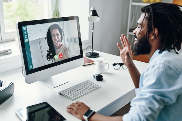 Confident young man in shirt talking with smiling woman using computer while sitting indoors