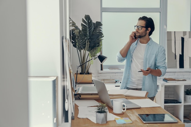 Confident young man talking on mobile phone while standing near his working place in office