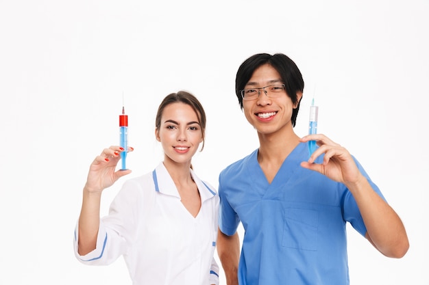 Confident young multiethnic doctors couple wearing uniform standing isolated over white wall, showing syringes