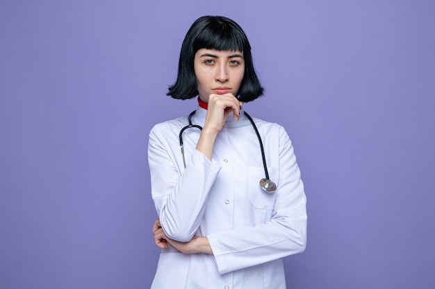 Confident young pretty caucasian girl in doctor uniform with stethoscope putting hand on her chin and looking at front