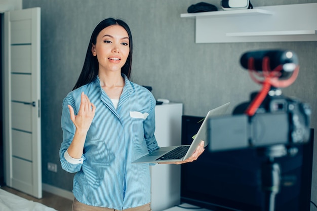 Confident young woman standing with a modern laptop and gesturing while talking on a camera at home