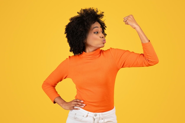 Confident young woman with afro hair flexing her arm muscle in a strong pose wearing an orange