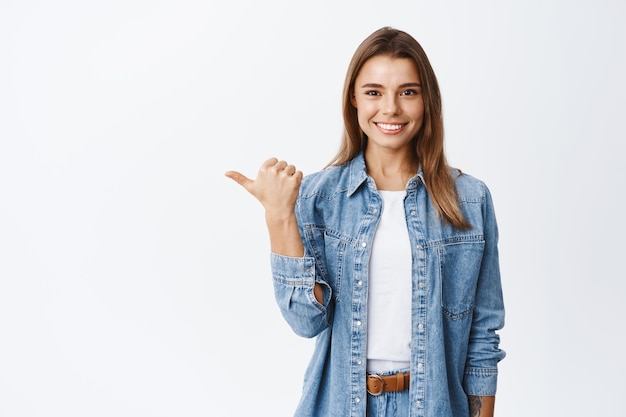 Confident young woman with assured smile pointing left at copy space logo, showing good deal, recommending product, white