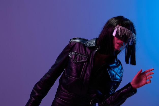 Confused adorable brunet woman in leather jacket specular sunglasses open mouth look aside posing isolated in blue violet color light background Neon party Cyberpunk concept Copy space Good offer