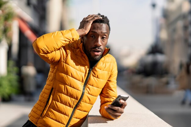 Confused African man with mobile phone complaining about mistake sitting in a city