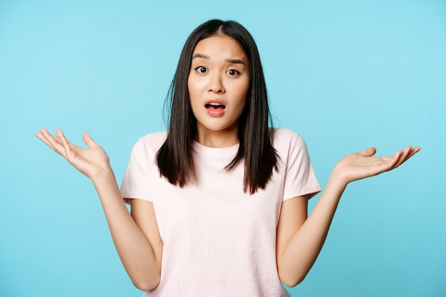 Confused asian girl spread empty hands sideways, shrugging shoulders clueless, standing in tshirt over blue background.