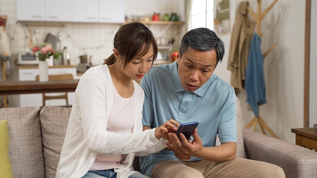 confused asian Japanese grandpa asking adult granddaughter questions while using cellphone in living room. the woman puts down her phone and teaches him how to use