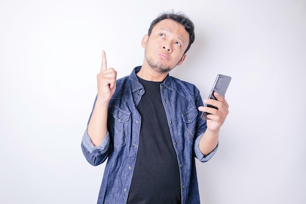 Confused Asian man wearing a navy blue shirt pointing at the copy space upside him holding his phone isolated by a white background