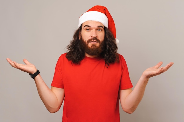 Confused bearded man is shrugging while wearing Christmas hat in a studio