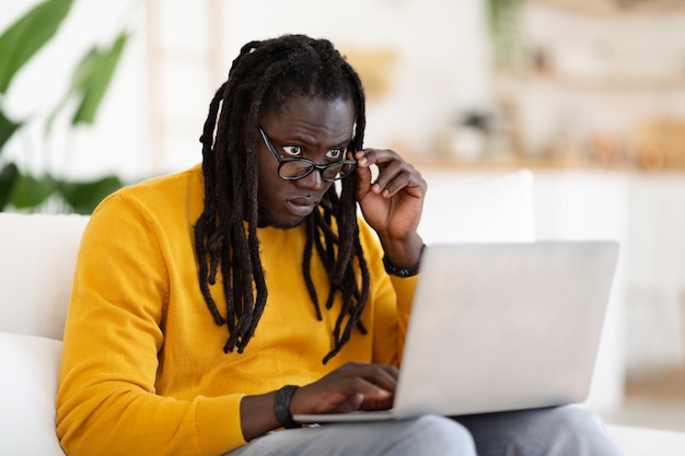 Confused Black Man Looking At Laptop Screen While Using Computer At Home