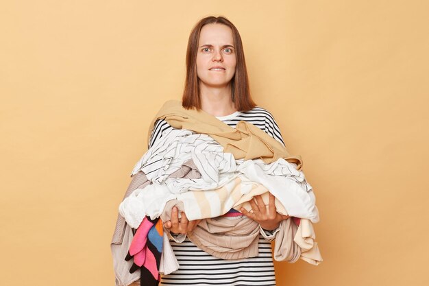 Photo confused brunette woman holding pile of clothes in hands isolated over beige background looking at camera with confused expression biting her lip