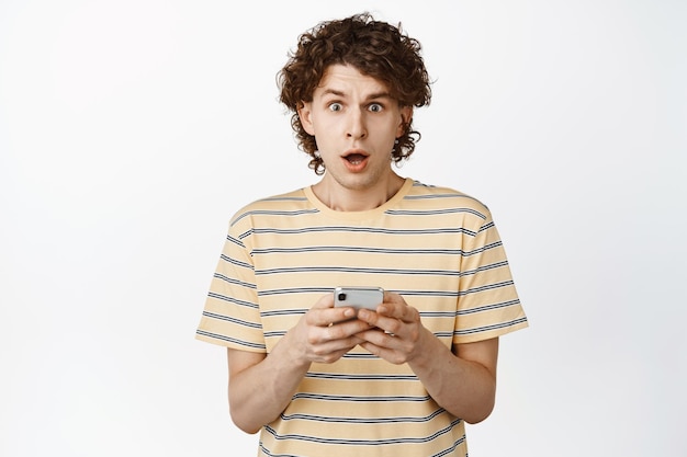 Confused curly guy looking at camera shocked after using mobile phone standing over white background copy space