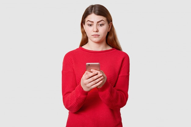 confused cute woman with dark straight hair, wears red sweater, holds mobile phone in hand, looking at its display, isolated on white. People amotion concept.