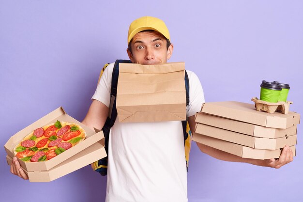 Confused delivery man wearing t shirt and cap holding pizza boxes and takeaway coffee standing with paper parcel in mouth looks helpless having lots orders posing isolated over purple background