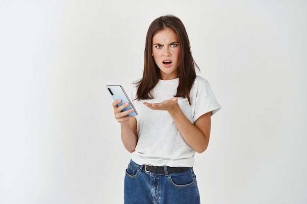 Confused and frustrated young woman having problem with smartphone, complaining on phone, standing over white wall