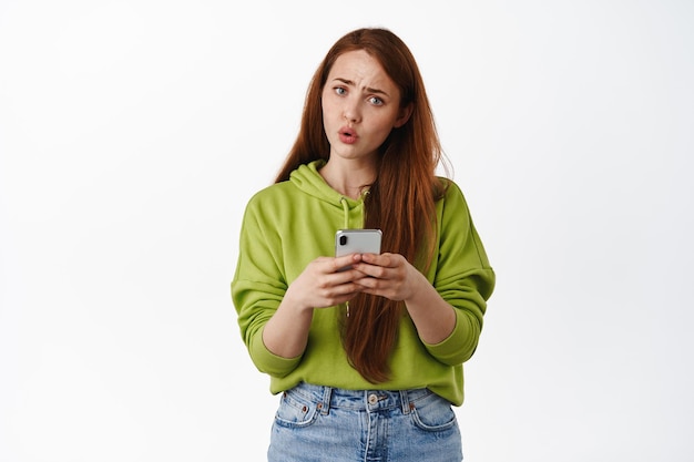 Confused ginger girl holding phone and stare puzzled, strange message on smartphone, cant understand how app works, standing over white background.