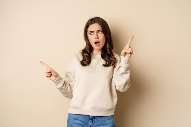 Confused girl shopper pointing sideways, making choice, decision between two variants, beige background