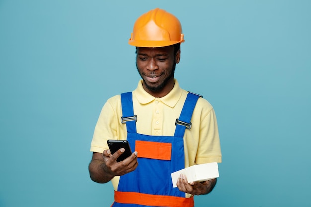 Confused holding brick and looking at phone in his hand young african american builder in uniform isolated on blue background