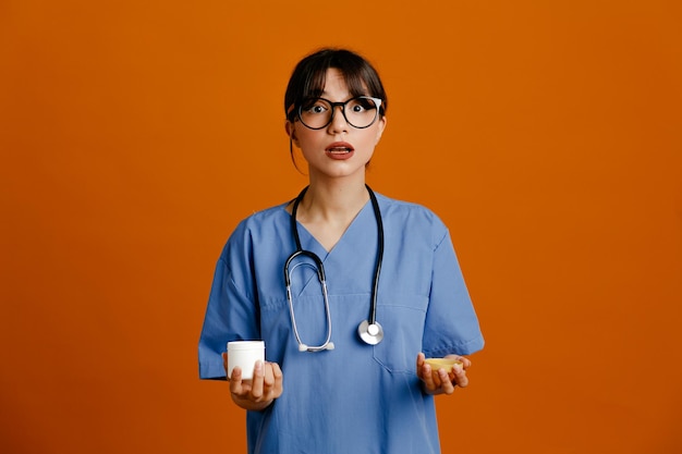 Confused holding can of pill young female doctor wearing uniform fith stethoscope isolated on orange background