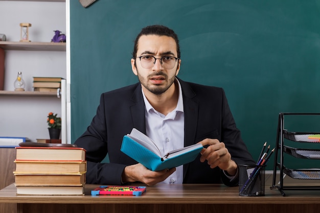 Confused male teacher wearing glasses holding book sitting at table with school tools in classroom