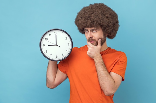 Confused man holding big wall clock and holding chin thinks about deadline