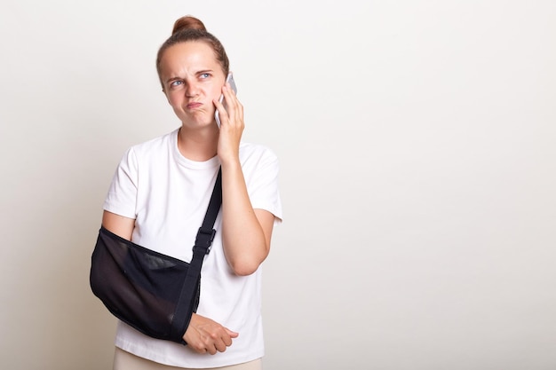 Confused pensive woman standing in arm sling after injuring hand talking on cell phone looking away having puzzled facial expression isolated on light gray background Copy space for advertisement