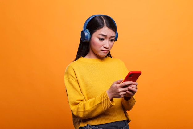 Confused puzzled woman feeling helpless because of faulty smartphone device. Doubtful young adult asian person wearing headphones while having mobile cellphone on orange background.