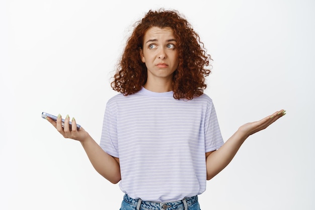 Photo confused redhead woman hold her mobile phone, shrugs and looks aside puzzled, dont know, cant understand smth perplexing, stands against white background